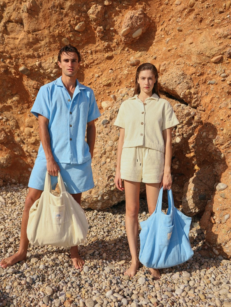 Man in coastal blue and woman in off-white organic cotton terry shirts and shorts holding matching beach bags by Little Mariner, standing on rocky beach.