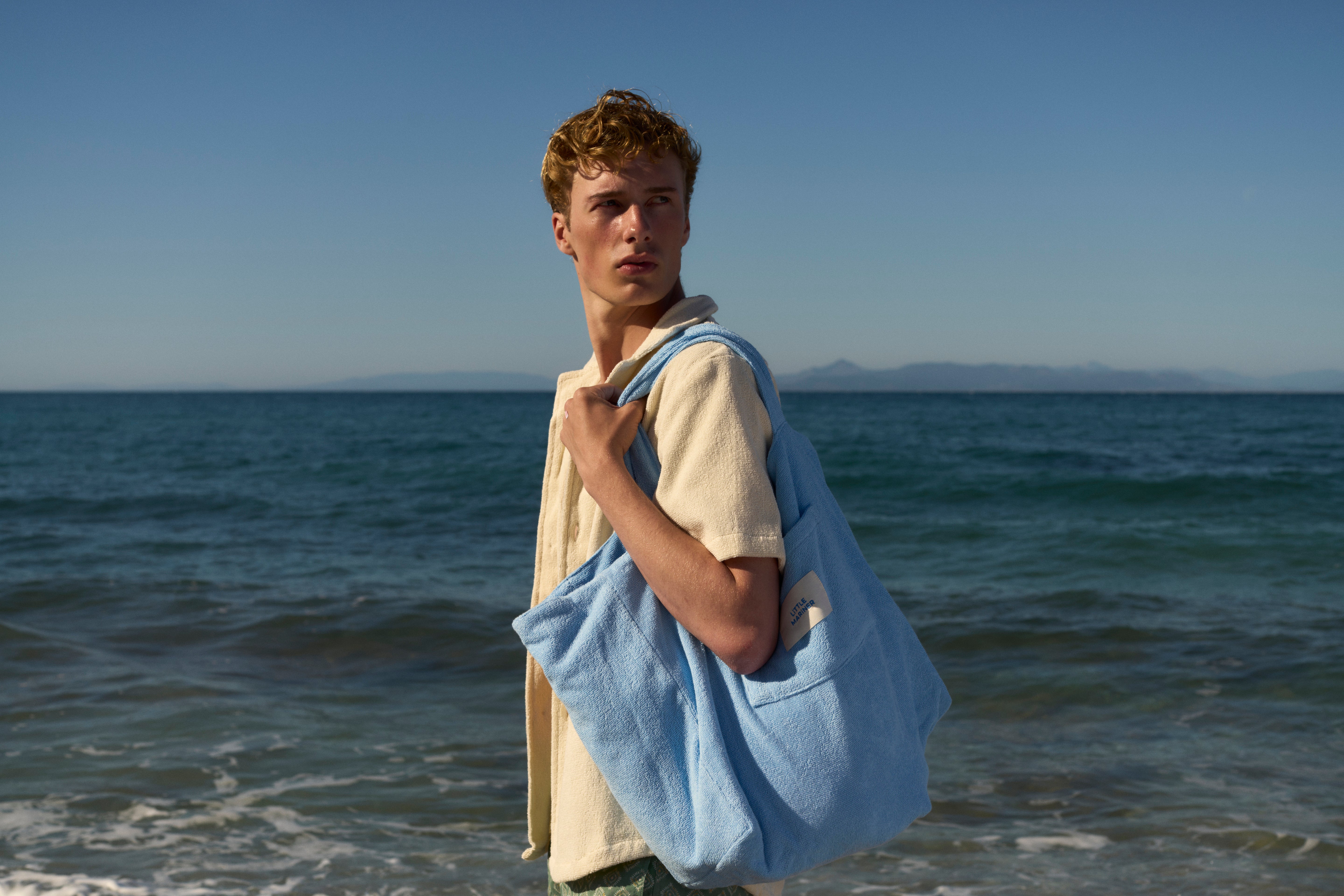 Man carrying a Coastal Blue organic cotton terry tote bag on his shoulder by the sea, in an Off-White terry shirt
