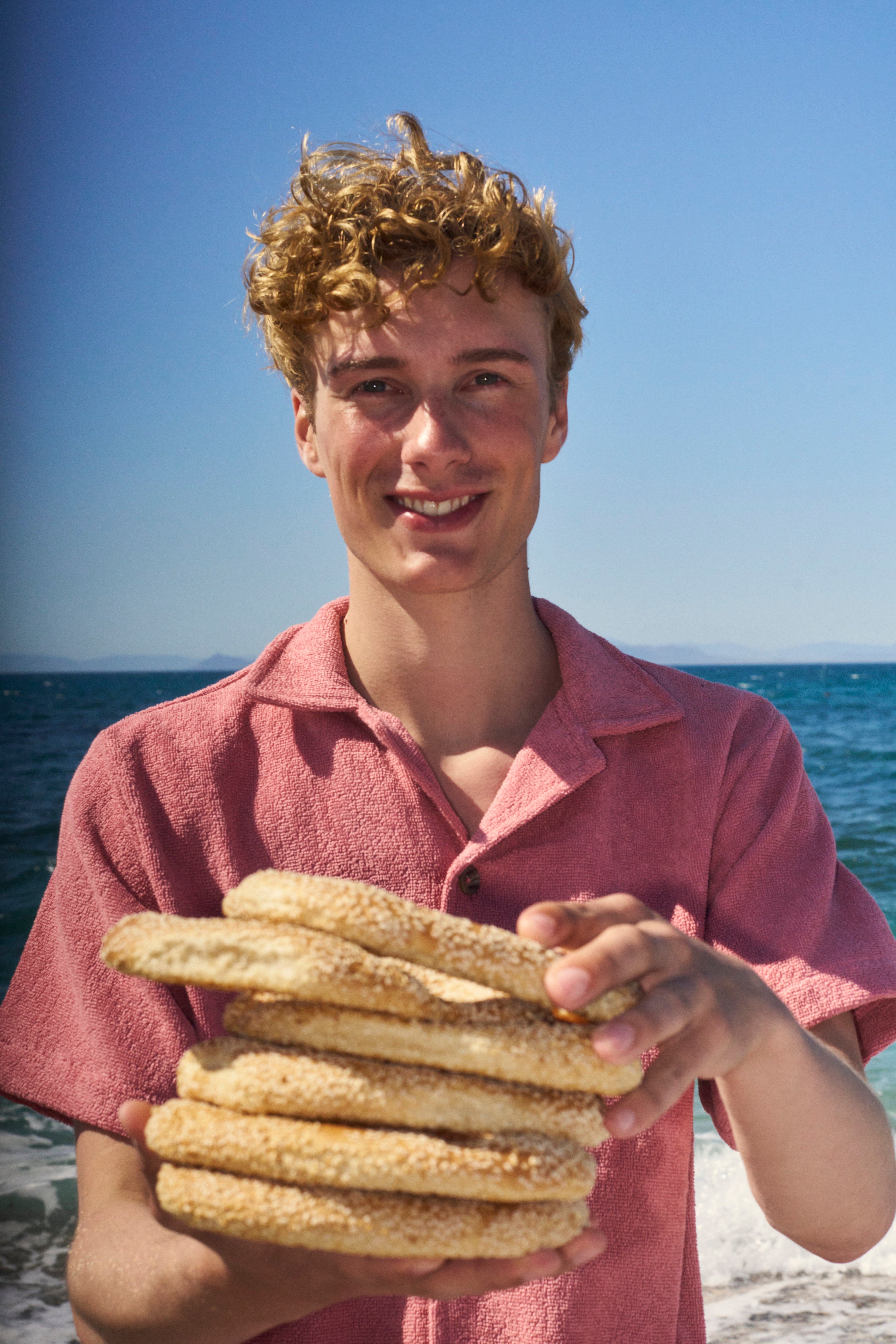 Smiling man in a Dusty Rose organic cotton terry camp-collar shirt holding sesame bread rings by the sea
