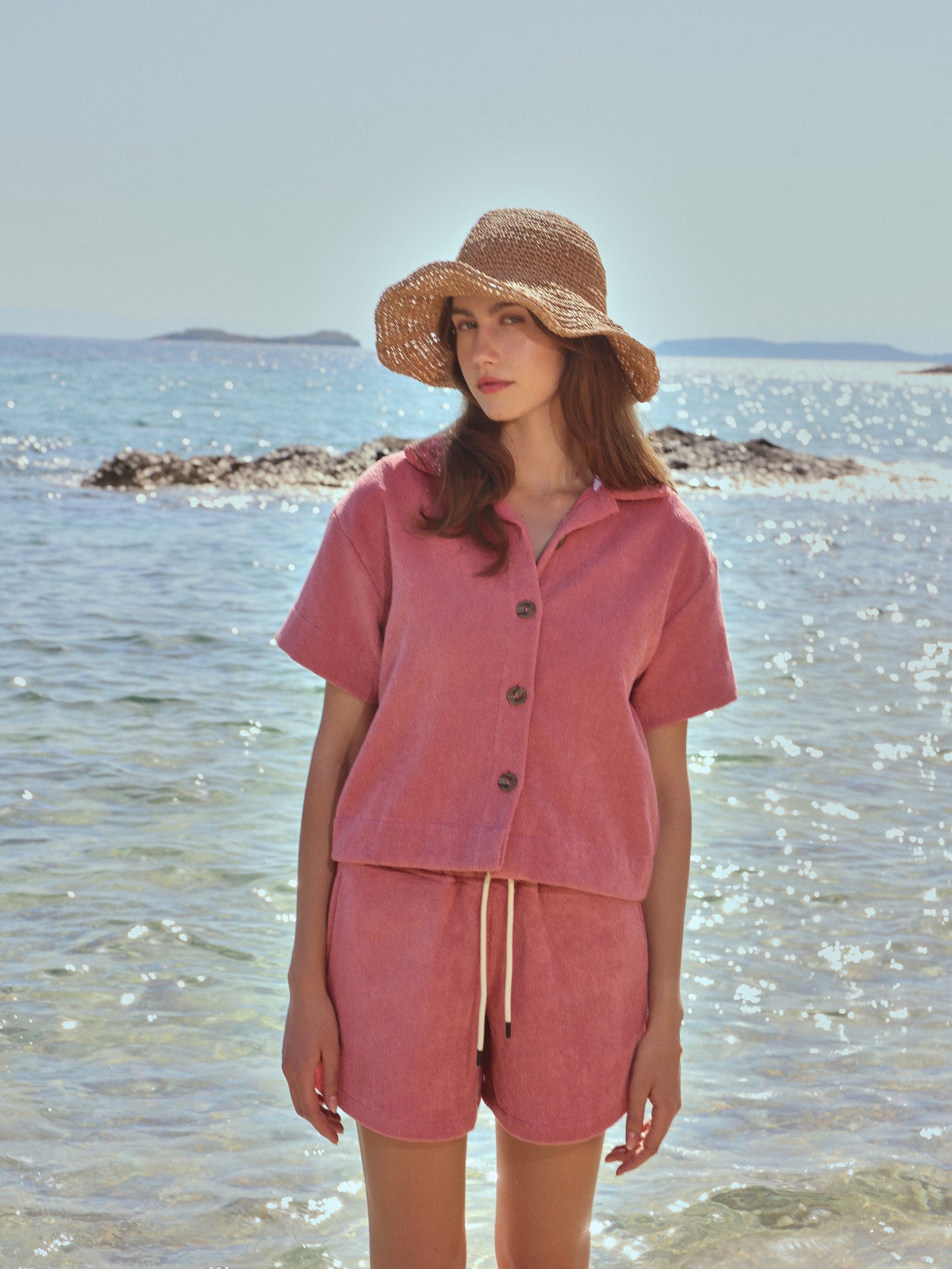 #Dusty Rose Woman in Little Mariner dusty rose terry shorts and shirt and straw hat standing on a beach with water and sky in the background