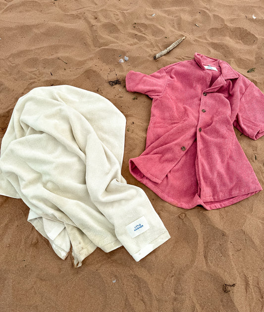Little Mariner off-white towel and dusty rose terry shirt on a sandy surface.