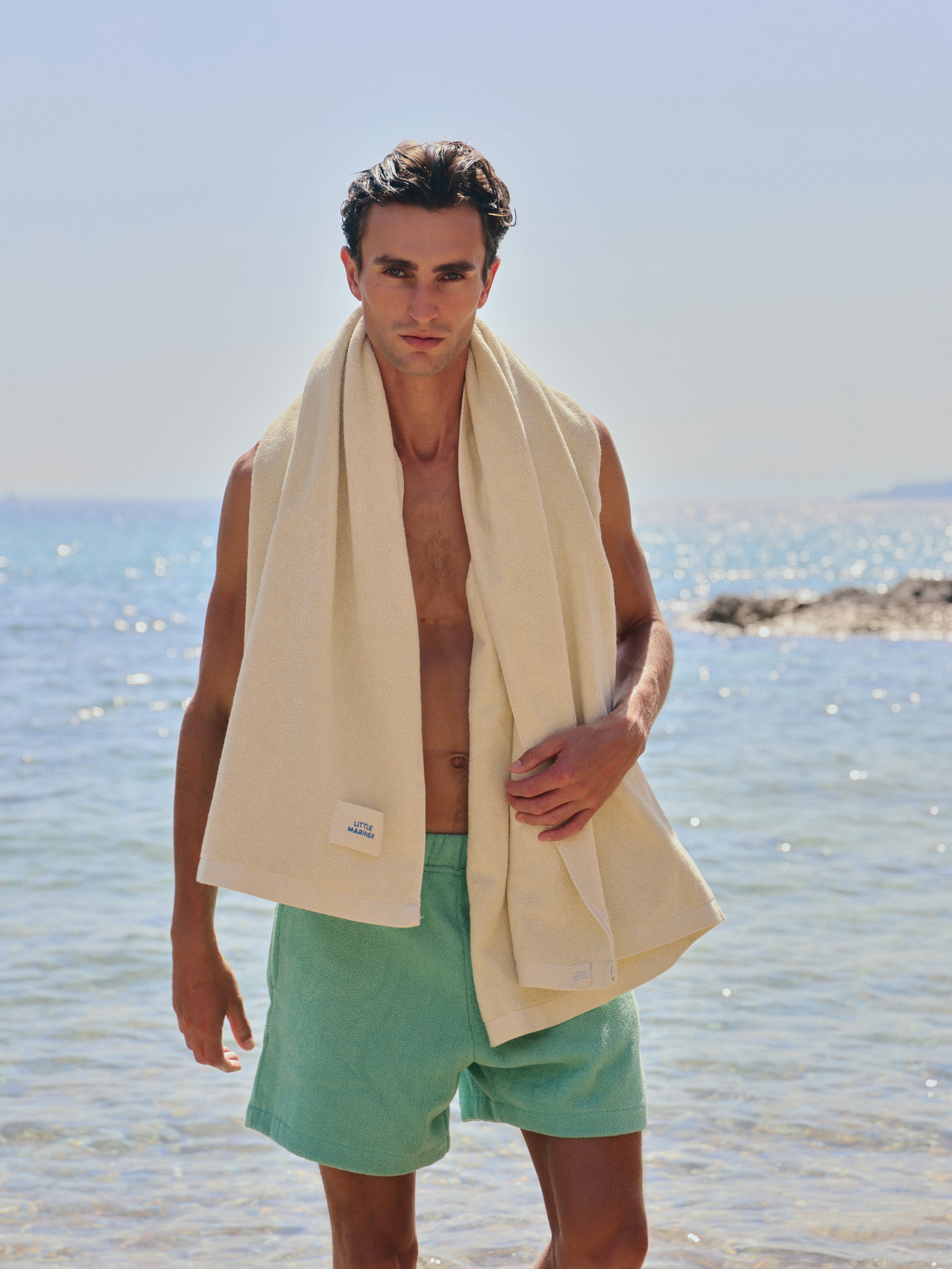#Sage Green Man standing on a beach wearing Little Mariner sage green terry shorts and a Little Mariner off-white towel draped over his shoulder.