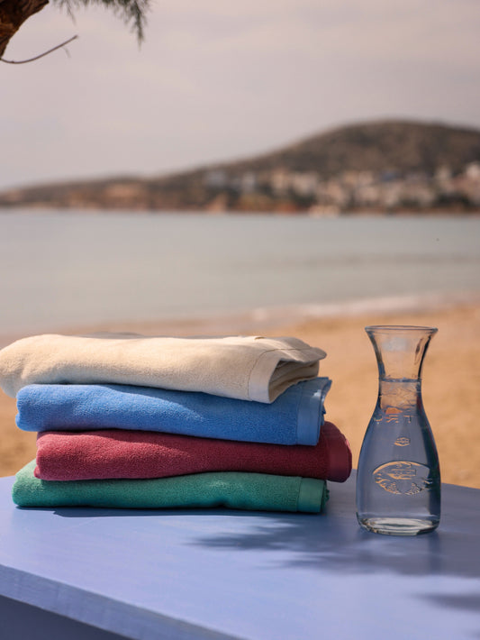 Stack of Little Mariner off-white, coastal blue, dusty rose and sage green towels with a glass carafe on a table by a scenic lakeside.