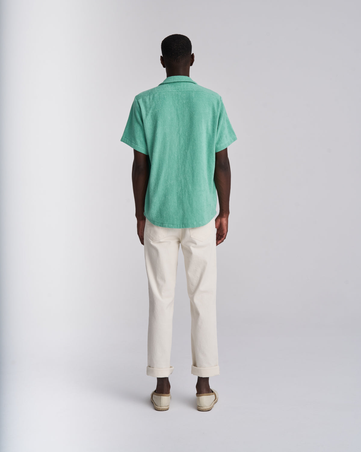 Back view of a man wearing a Sage Green organic cotton terry camp-collar shirt, buttoned, with cream trousers and espadrilles