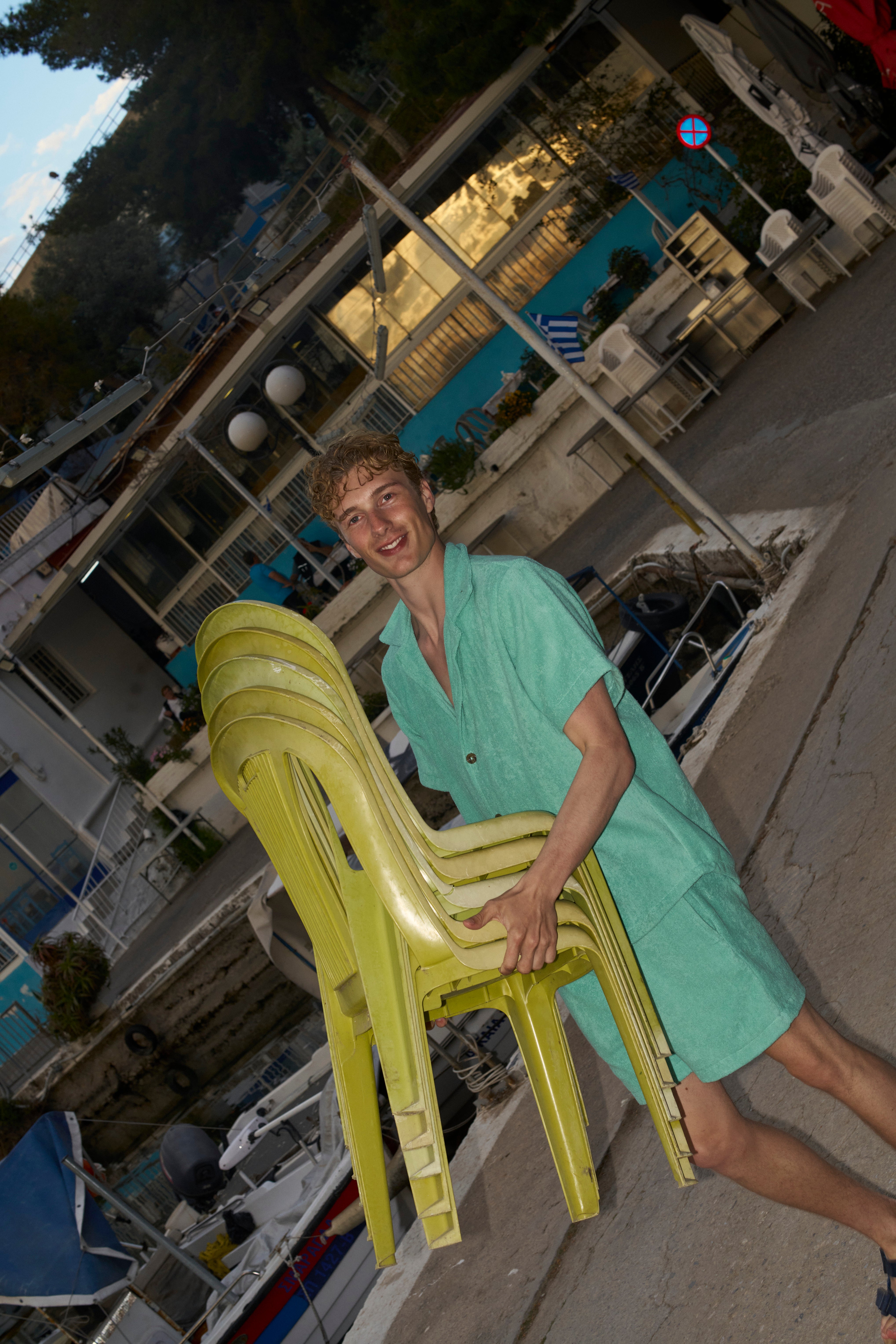 Man carrying stacked chairs in a Sage Green organic cotton terry shirt and matching shorts by the boats