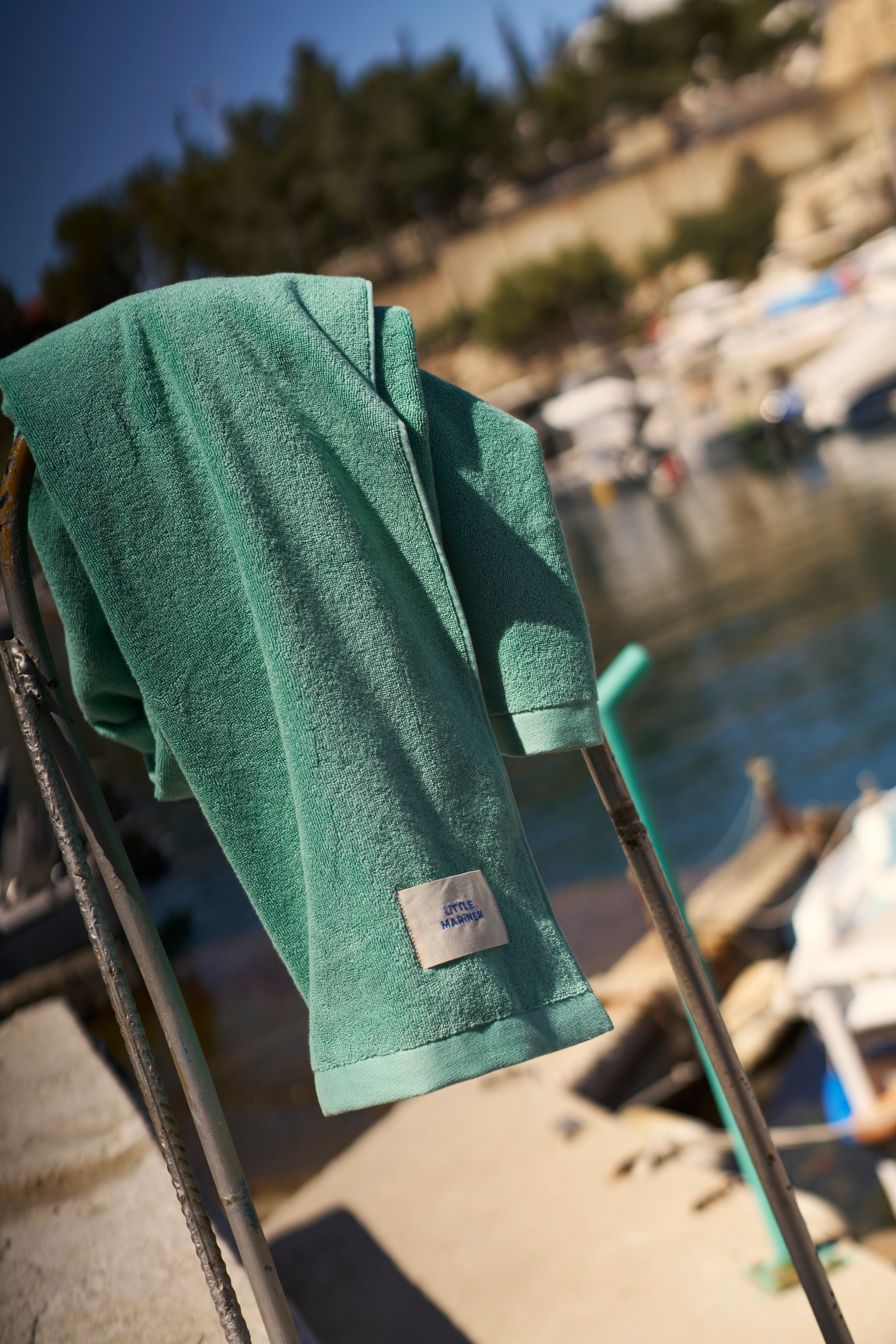 Sage Green organic cotton terry towel hung on a rail by a harbour, woven LiTTLE MARiNER label