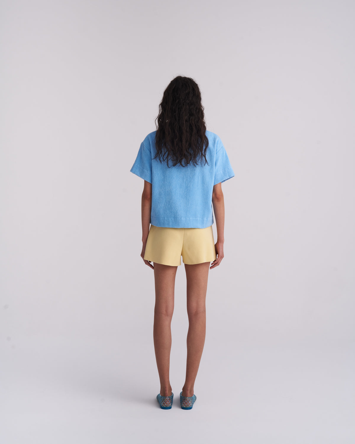 Woman in a Coastal Blue organic cotton terry camp-collar shirt with light yellow shorts, back view