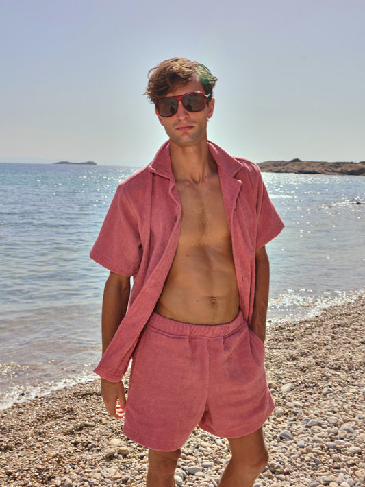 Man on rocky beach wearing dusty rose organic cotton terry shirt and shorts by Little Mariner, styled as open beachwear set.