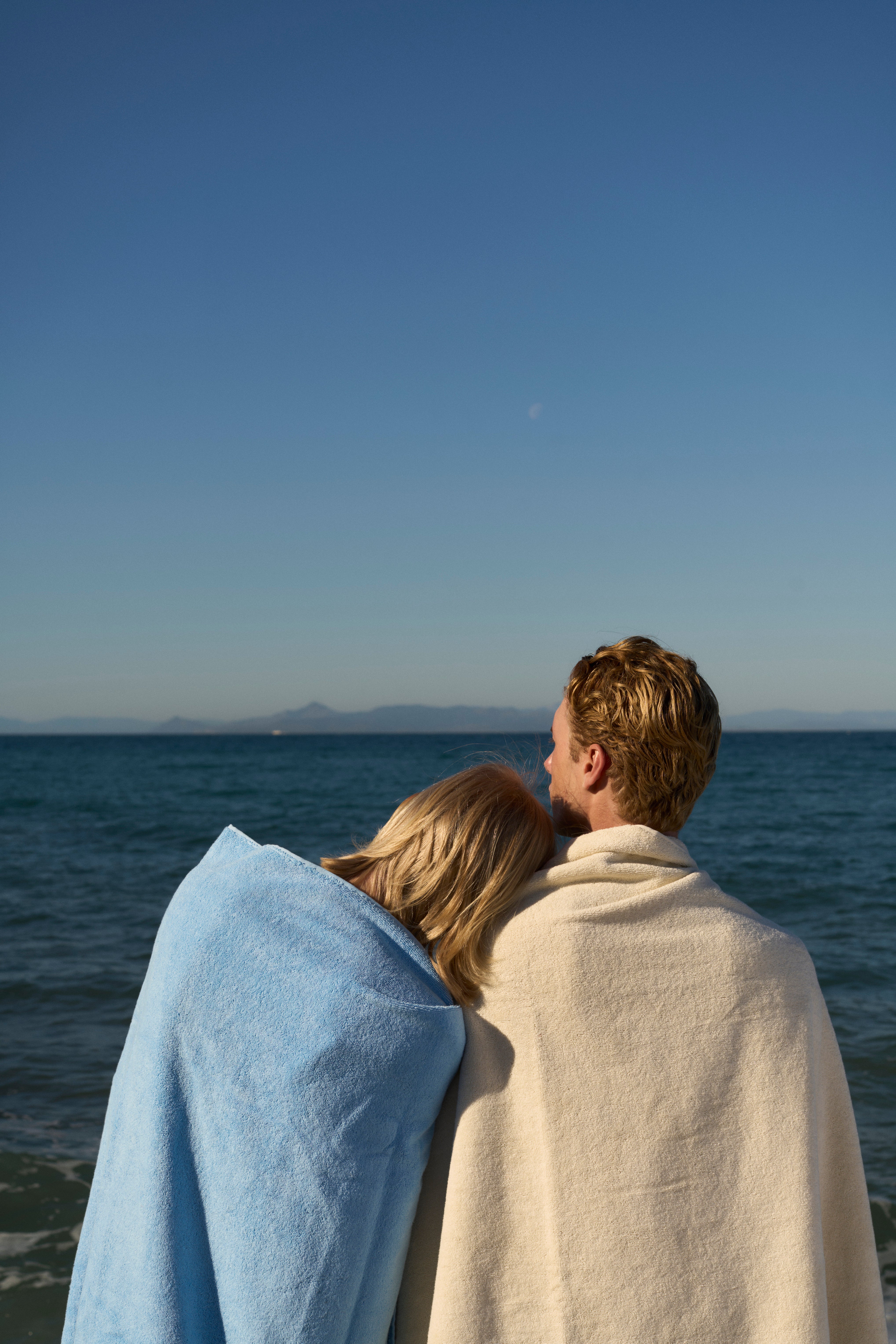 Couple wrapped in Coastal Blue and Off-White organic cotton terry towels by the sea