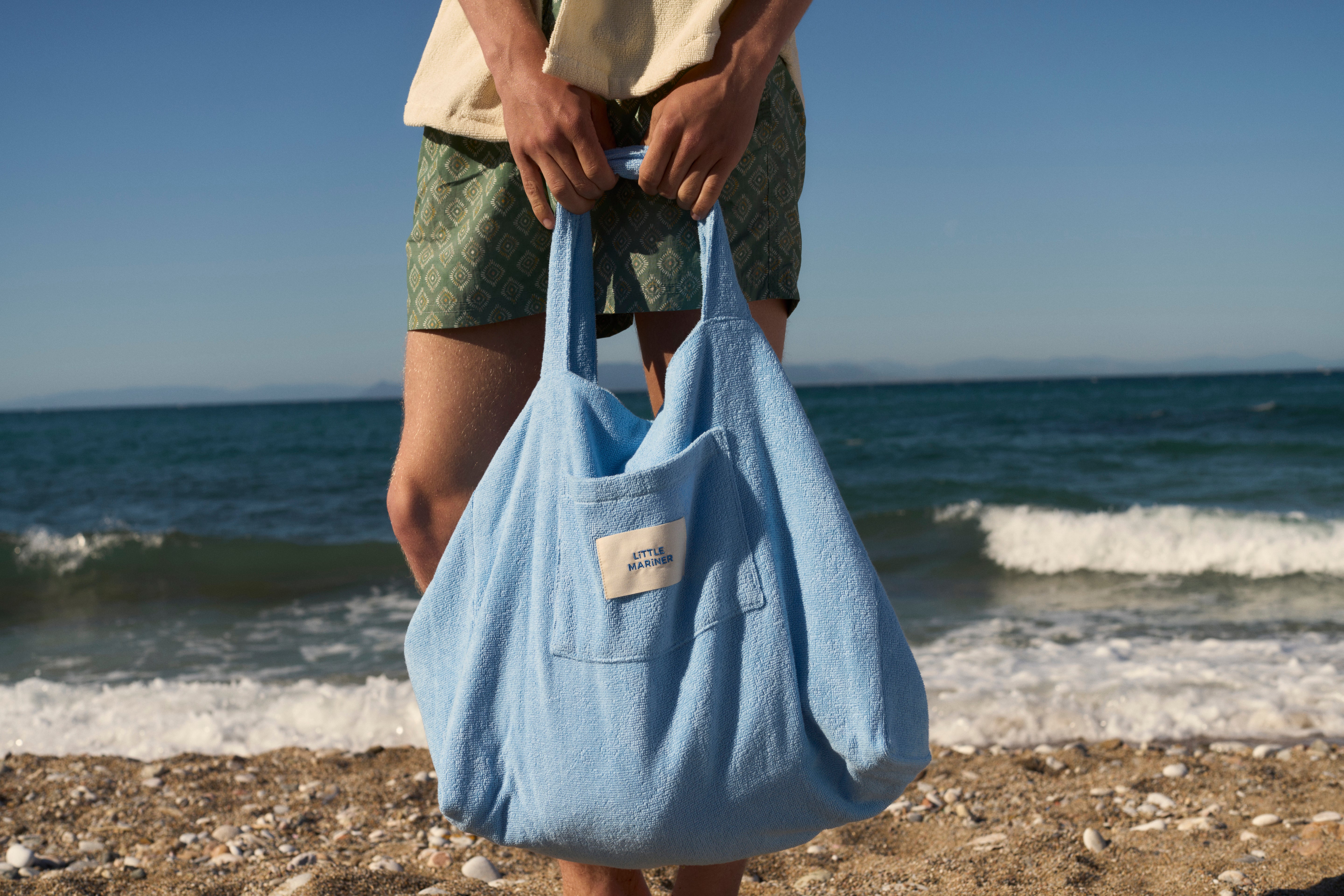 Coastal Blue organic cotton terry tote bag held by the handles on a beach, front pocket and woven label