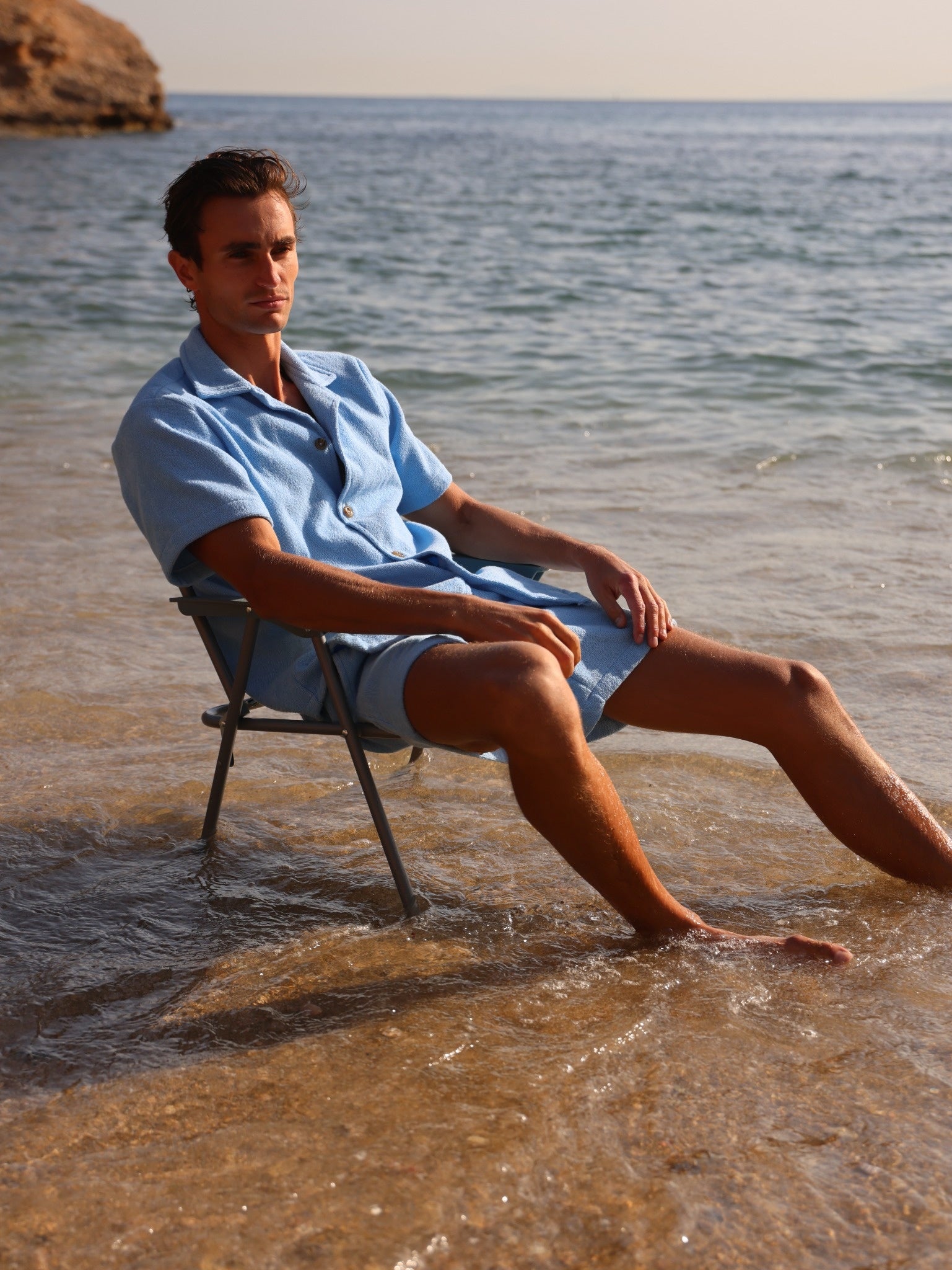 Man wearing Little Mariner coastal blue terry shirt and shorts sitting on a chair in shallow water at the beach