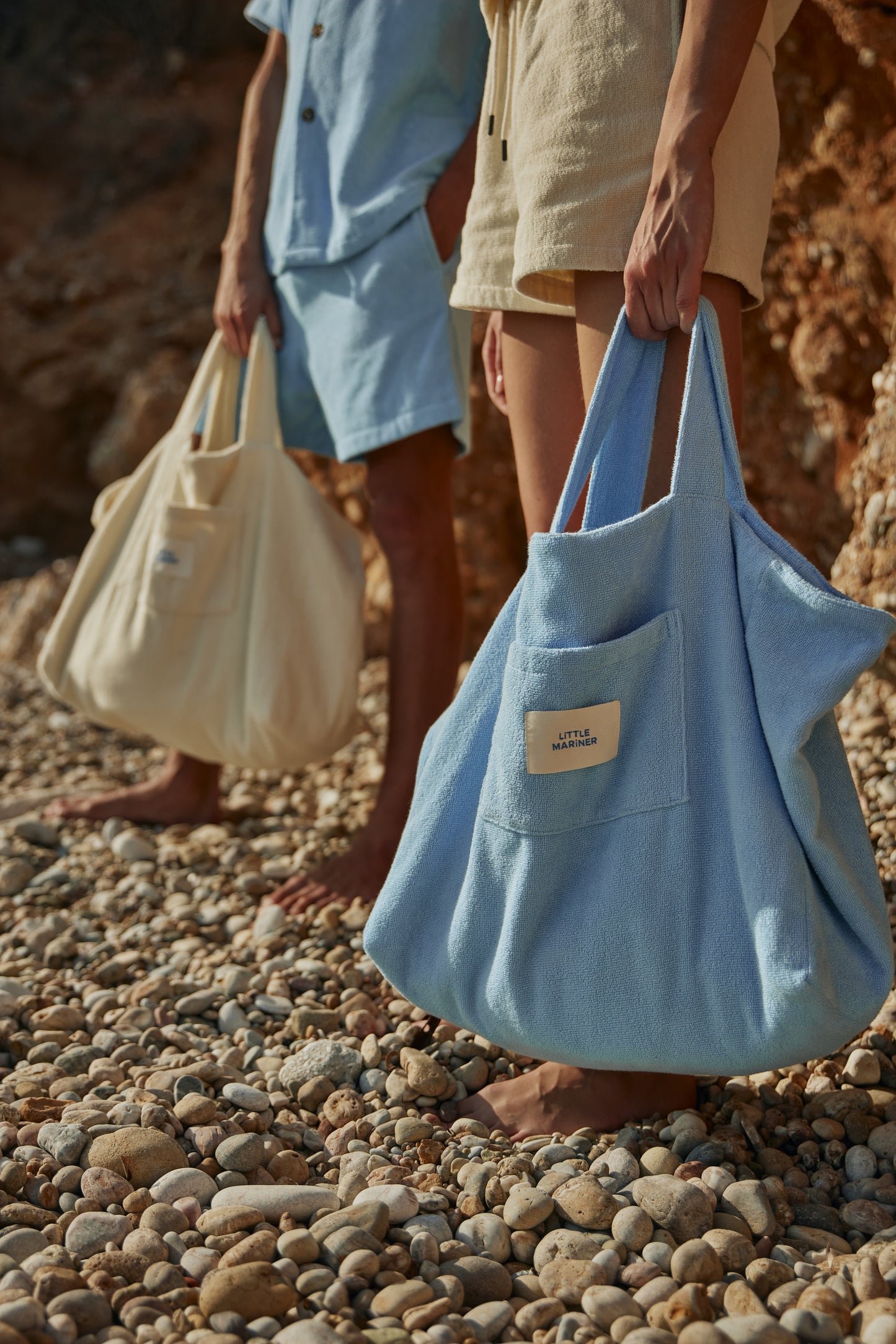 Two people holding Little Mariner off-white terry tote bags on a pebbly beach.