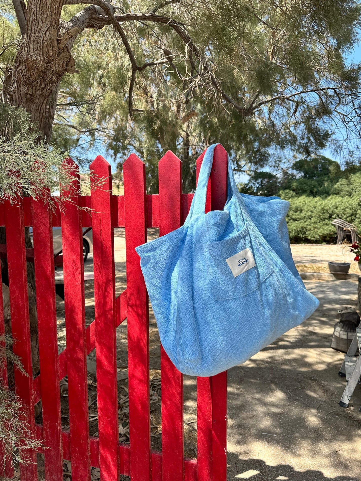 Little Mariner coastal blue bag hanging on a red wooden fence with trees and a person in the background