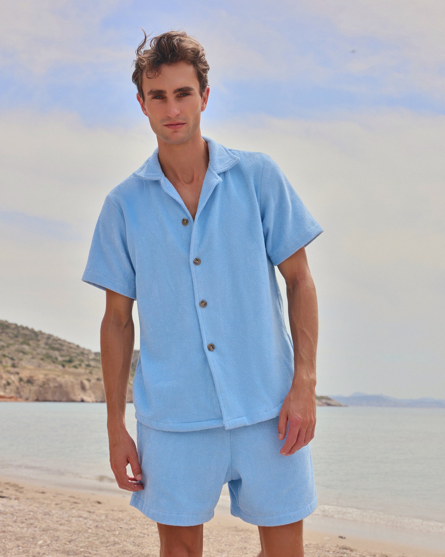 Man standing on beach wearing coastal blue organic cotton terry shirt and shorts set by Little Mariner.