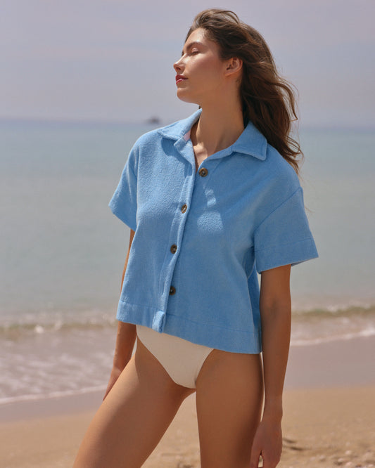 Woman on beach wearing coastal blue organic cotton terry short-sleeve shirt by Little Mariner, styled over a cream swimsuit.