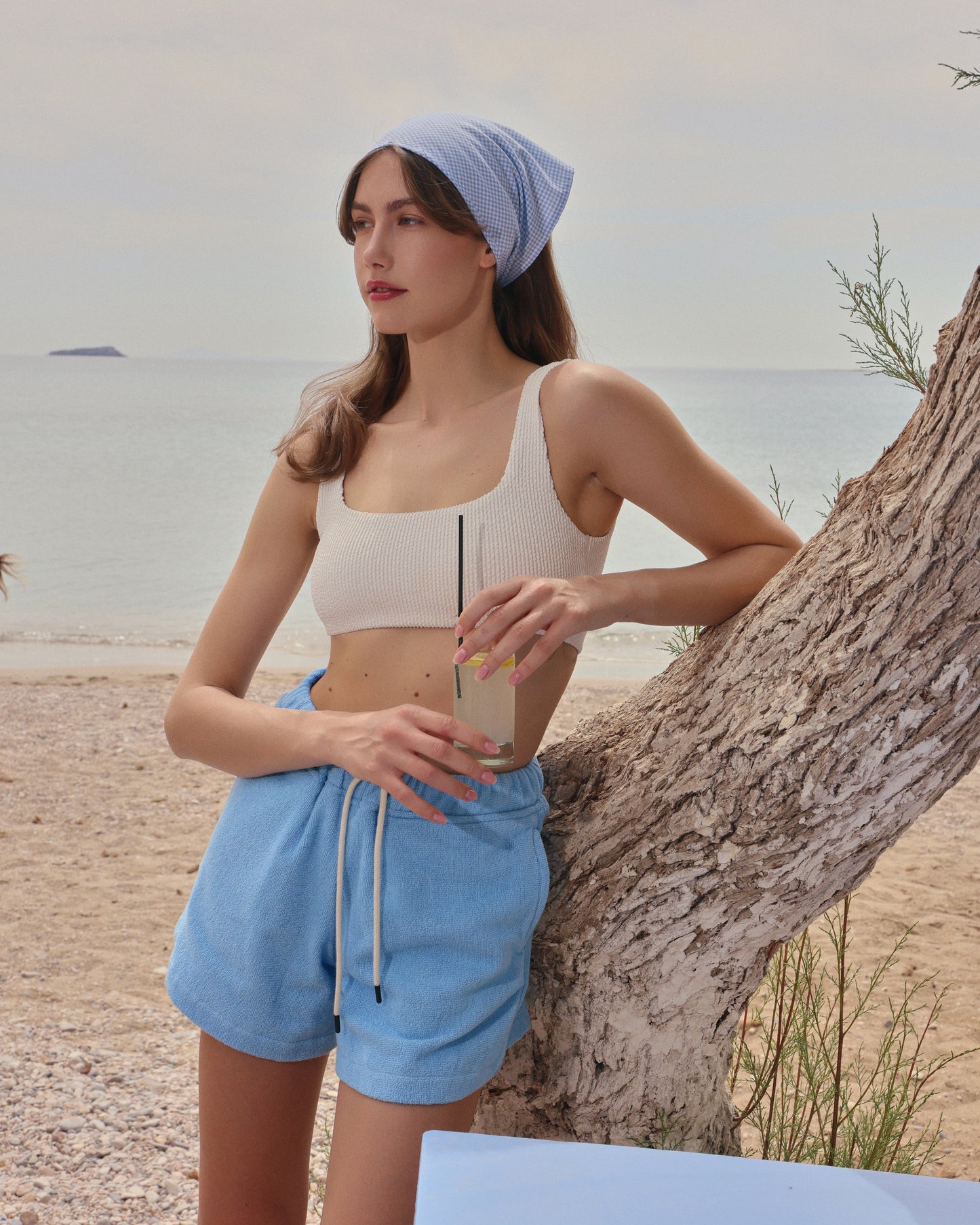 Woman on beach wearing coastal blue organic cotton terry drawstring shorts by Little Mariner, styled with a ribbed crop top and headscarf.