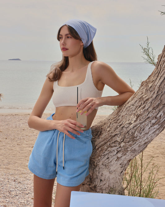 Woman on beach wearing coastal blue organic cotton terry drawstring shorts by Little Mariner, styled with a ribbed crop top and headscarf.