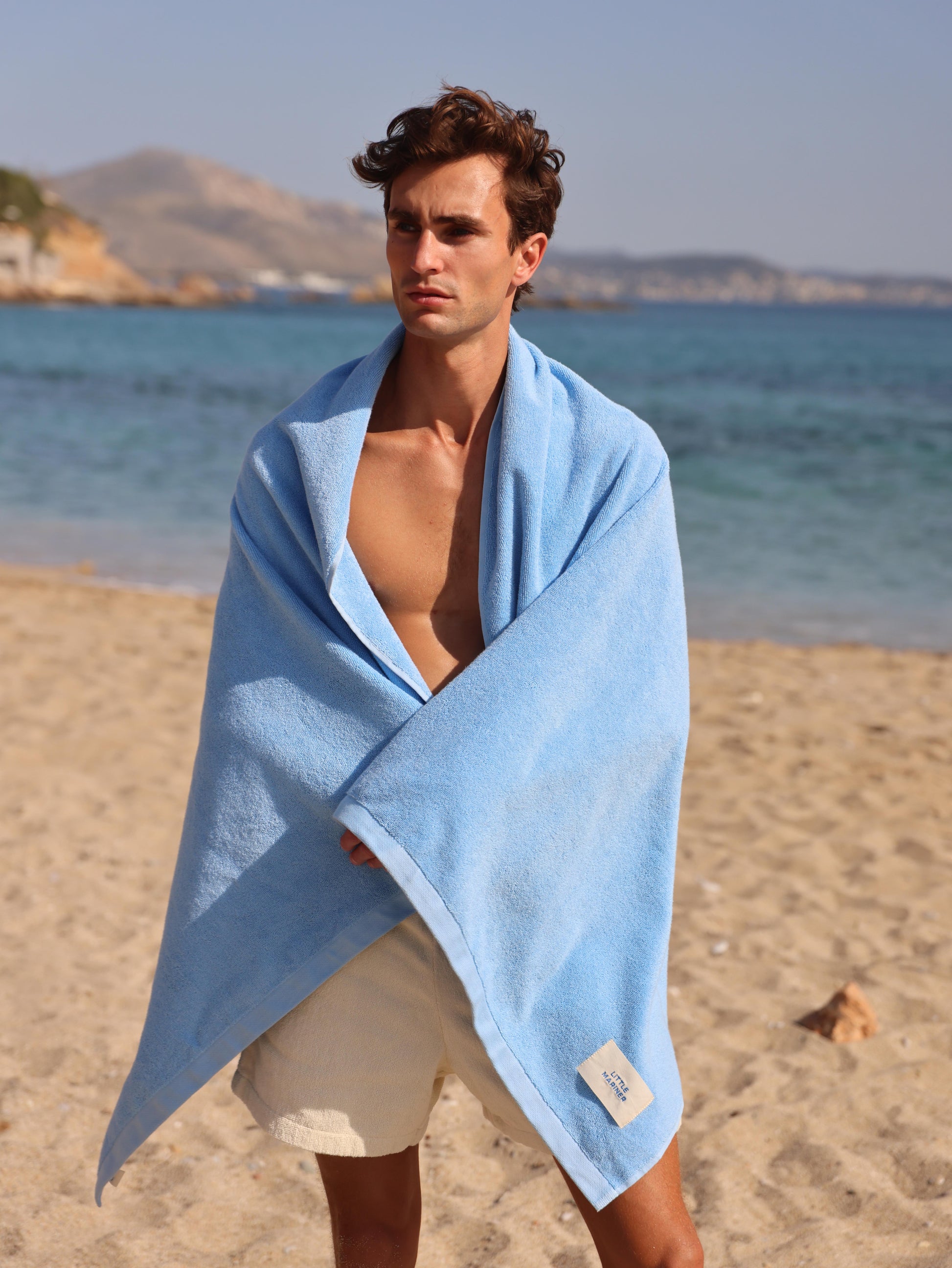Man wrapped in Little Mariner coastal blue towel on a beach with ocean and mountains in the background