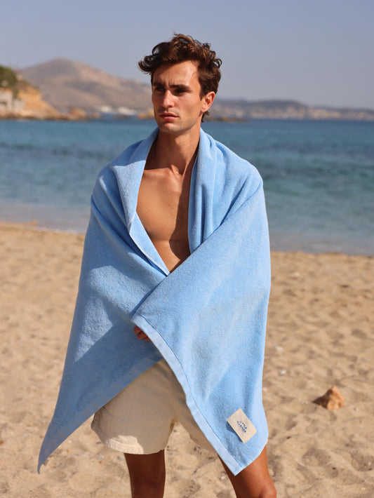 Man wrapped in Little Mariner coastal blue towel on a beach with ocean and mountains in the background