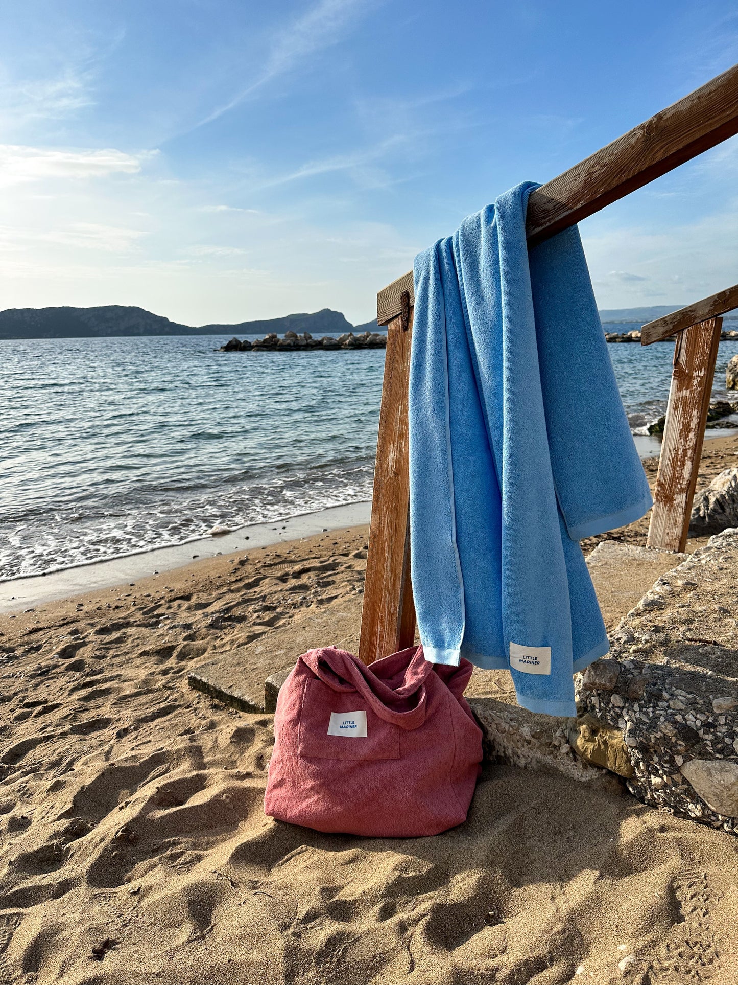 Little Mariner coastal blue towel and Little Mariner dusty rose terry bag on a beach with ocean and sky in the background