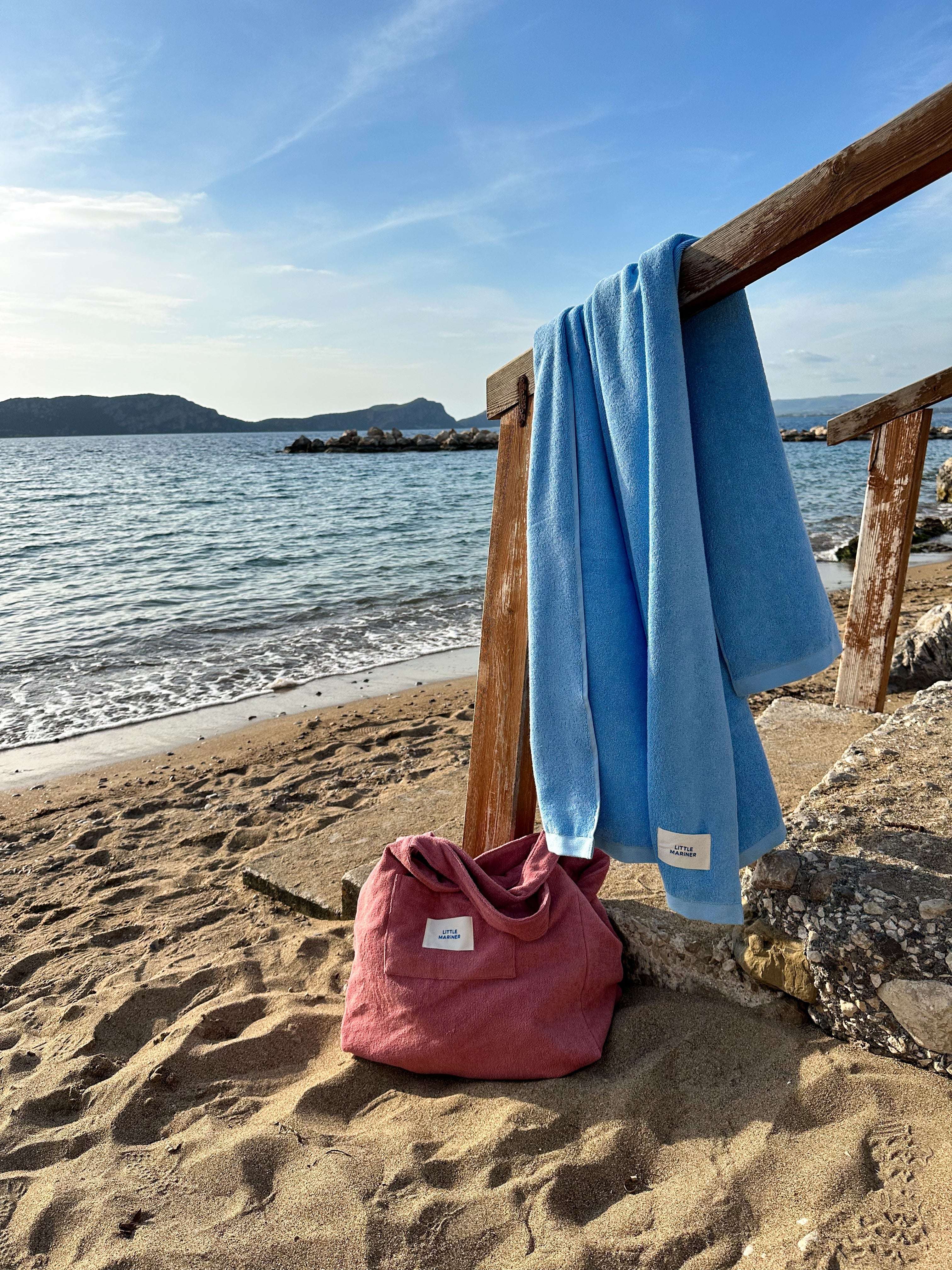 Little Mariner coastal blue towel and Little Mariner dusty rose terry bag on a beach with ocean and sky in the background