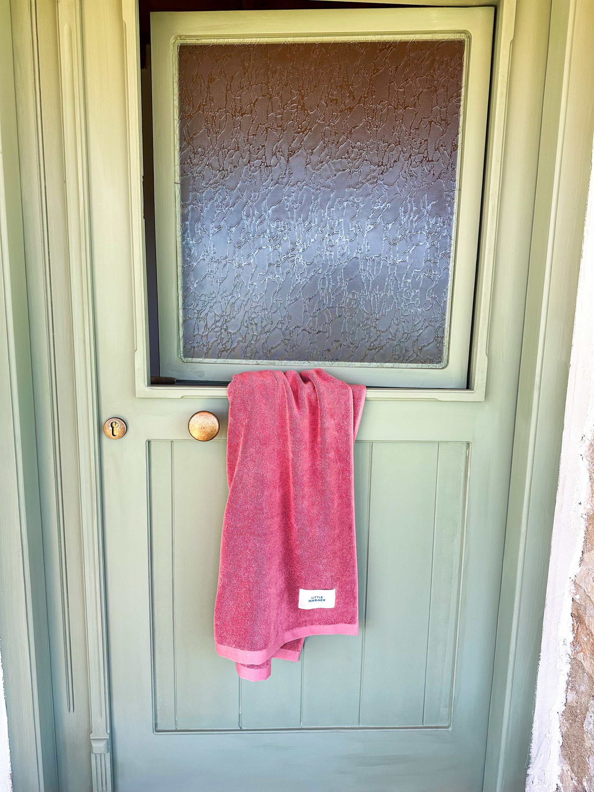 Little Mariner dusty rose towel hanging on a green door with a frosted glass panel