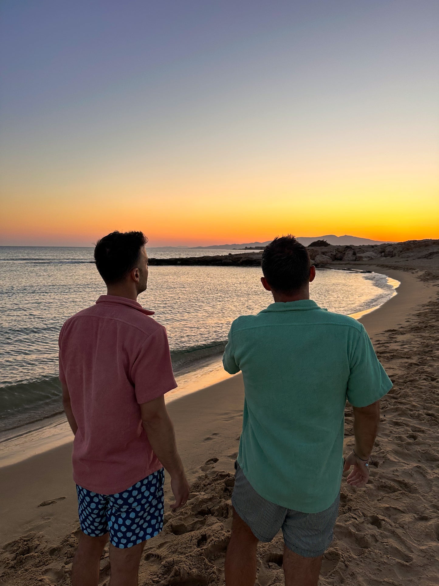 Two people wearing Little Mariner sage green and dusty rose terry shirts, standing on a beach at sunset, facing the ocean.