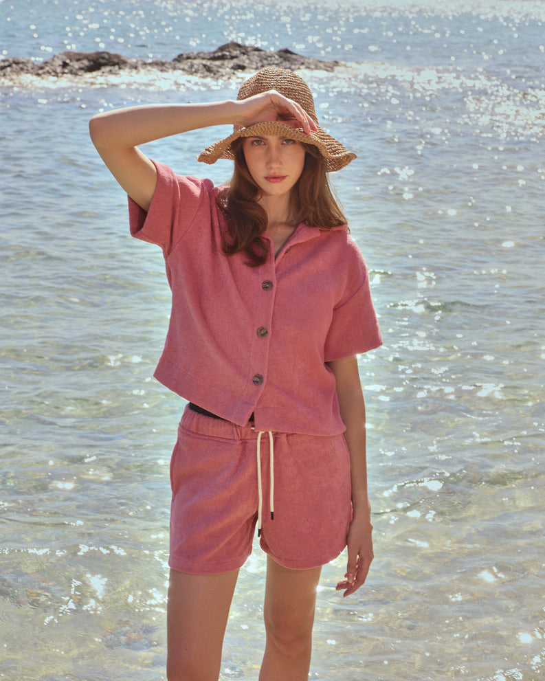 Woman on beach wearing dusty rose organic cotton terry shirt and drawstring shorts set by Little Mariner, styled with a straw hat.