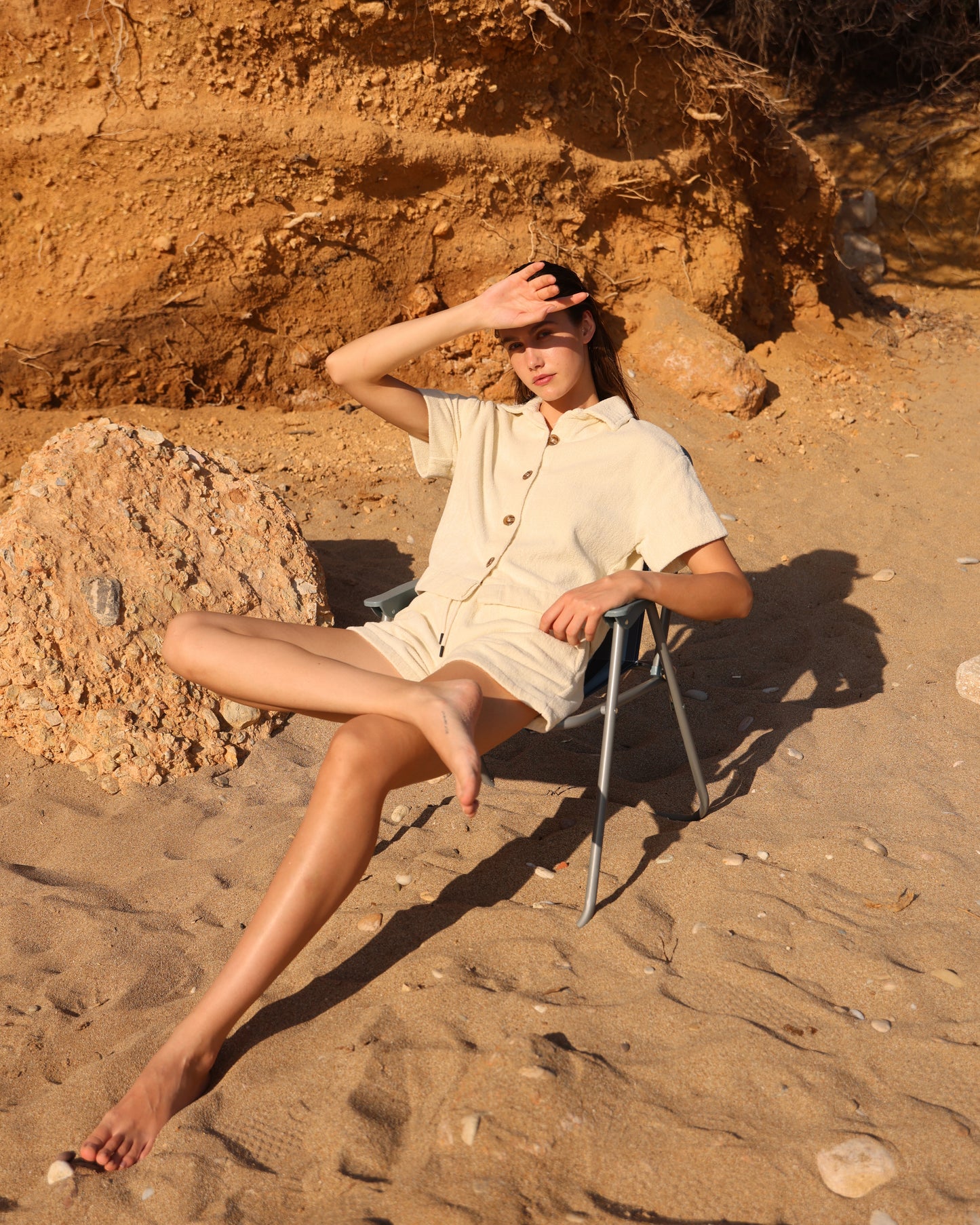 Woman sitting on a chair in Little Mariner off-white terry shirt and terry shorts setting with sand and rocky terrain
