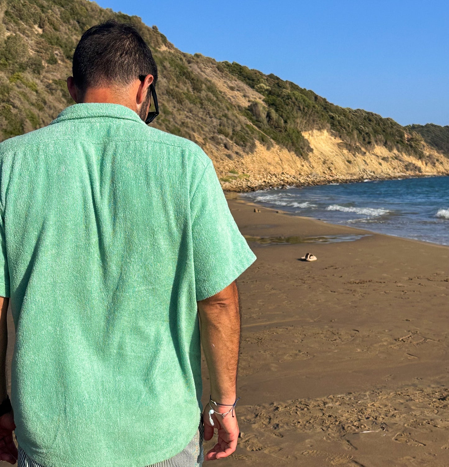 Person in Little Mariner sage green terry shirt standing on a beach with a scenic view of the ocean and hills.