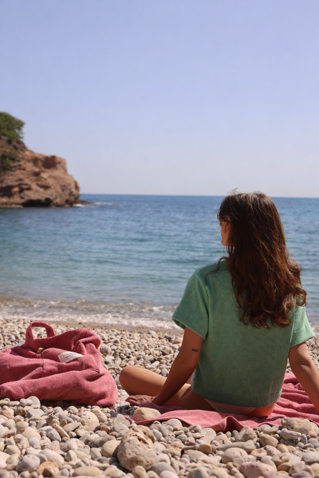 Woman wearing Little Mariner sage green terry shirt, sitting on Little Mariner dusty rose towel with Little Mariner dusty rose terry bag on a pebbly beach with ocean view