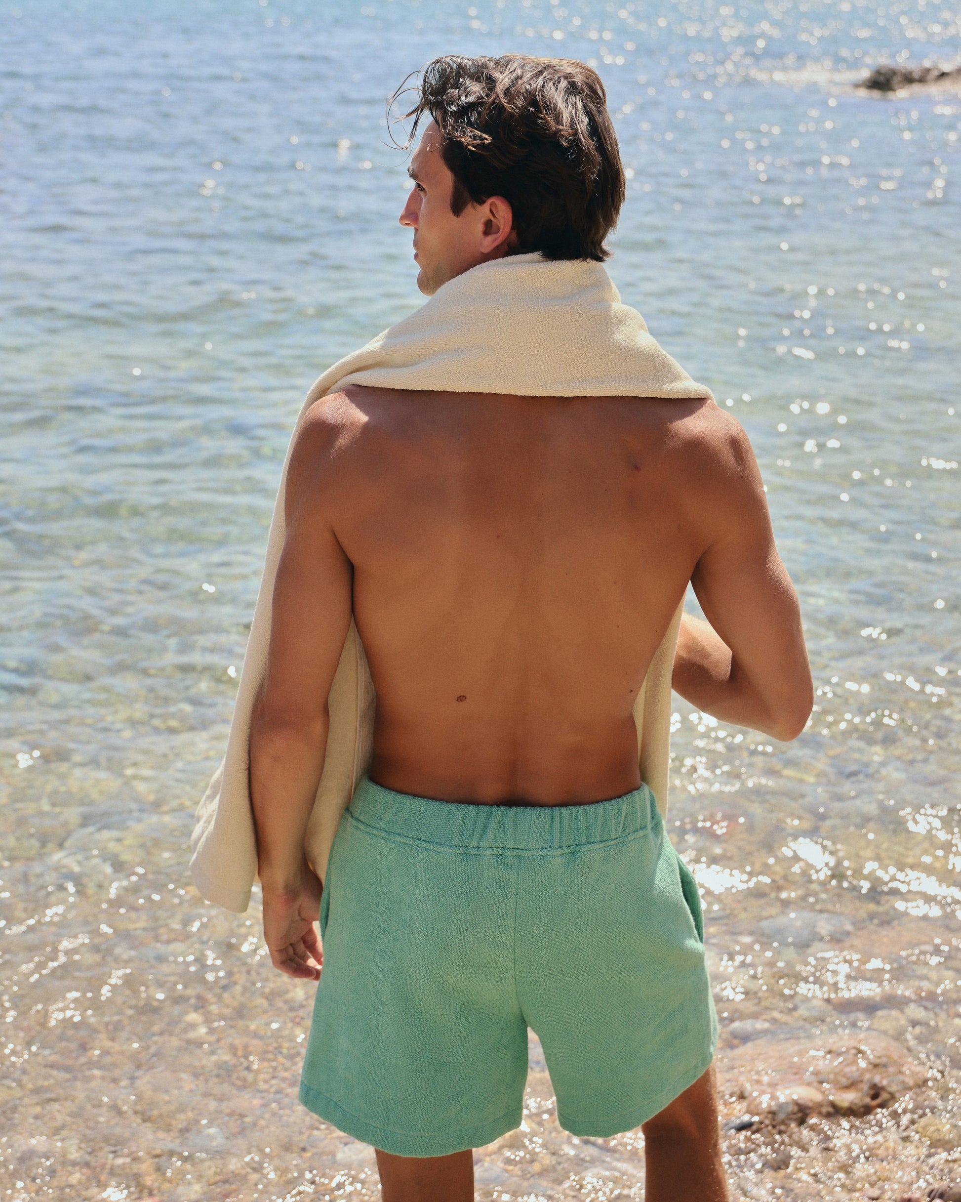Man standing on a beach wearing Little Mariner sage green terry shorts and a Little Mariner off-white towel draped over his shoulder.