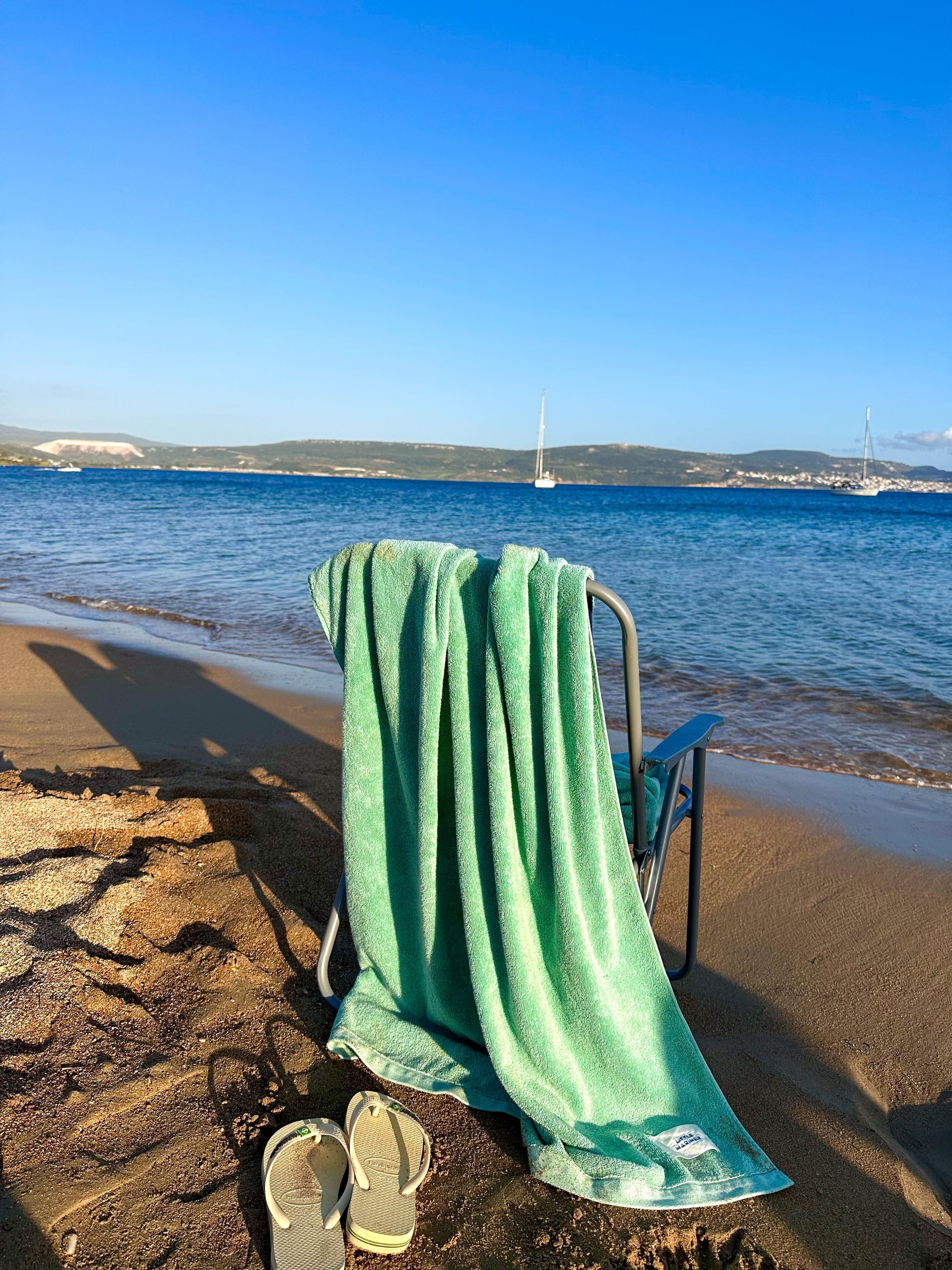 Beach chair with Little Mariner sage green towel draped over it on a sandy beach with ocean and clear sky in the background.