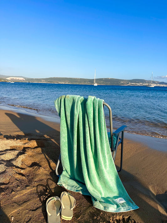 Beach chair with Little Mariner sage green towel draped over it on a sandy beach with ocean and clear sky in the background.