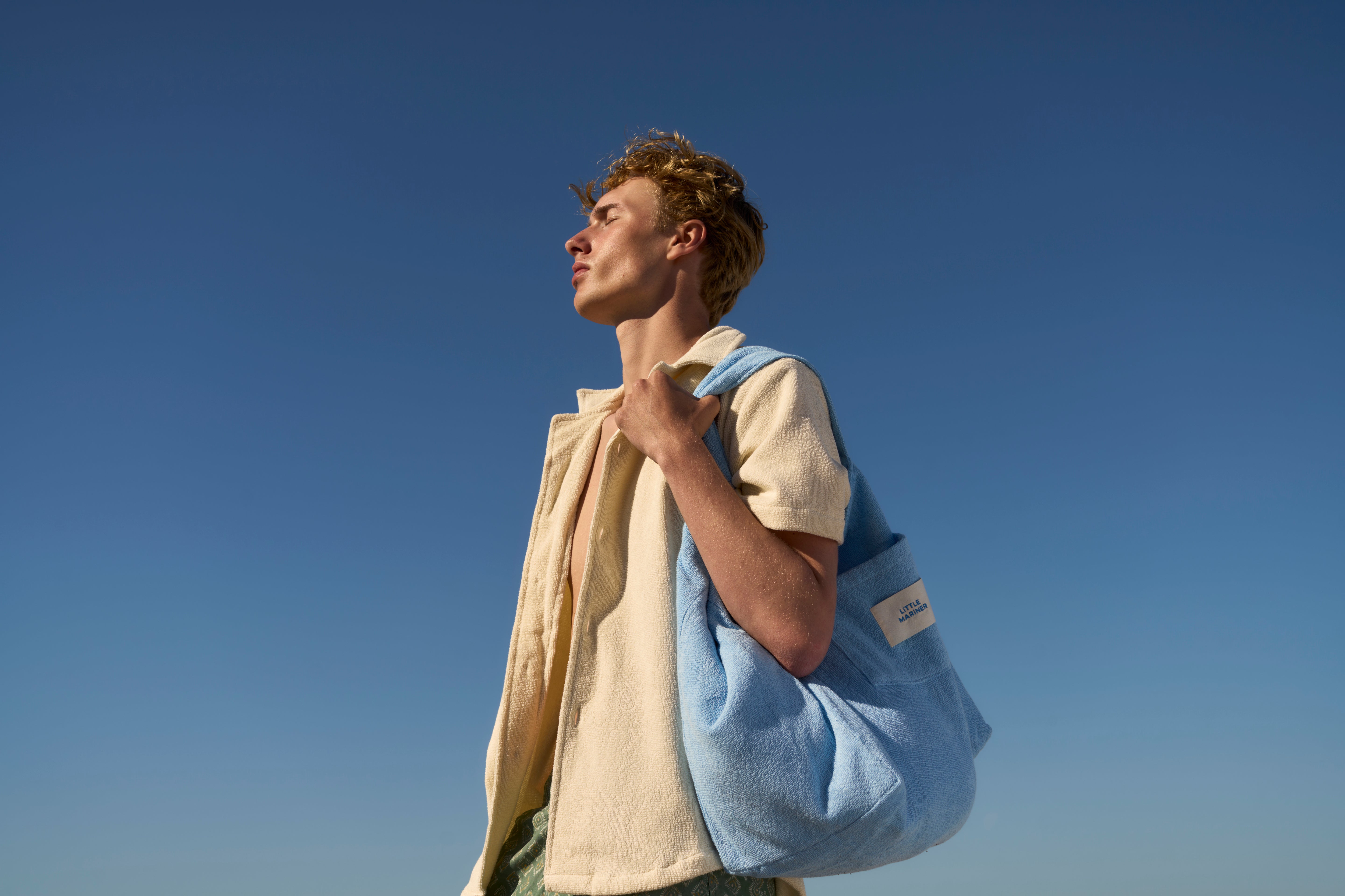 Man wearing an Off-White organic cotton terry shirt with a Coastal Blue terry tote bag, against the sky