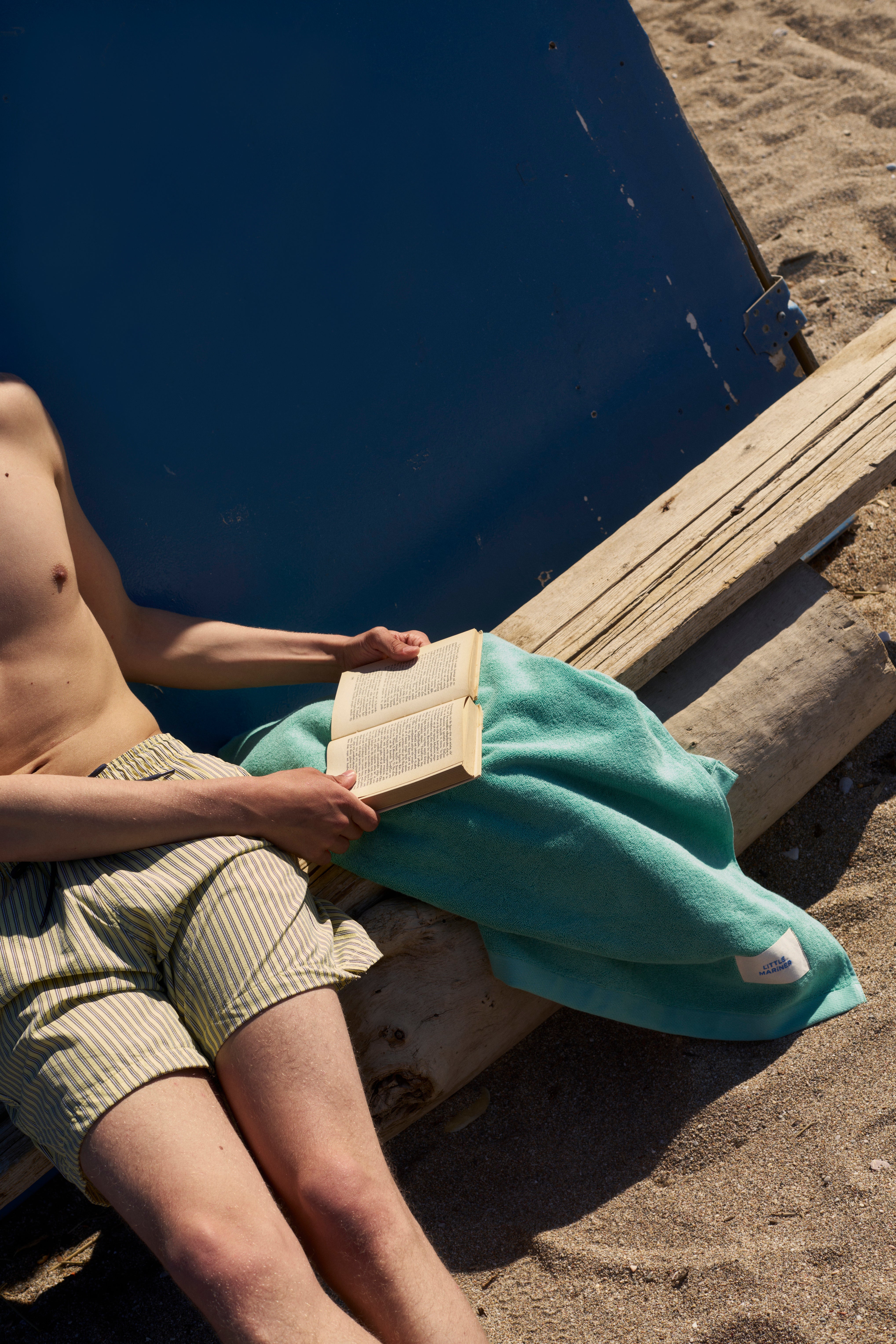 Man on a beach reading, with a Sage Green organic cotton terry towel draped over his lap