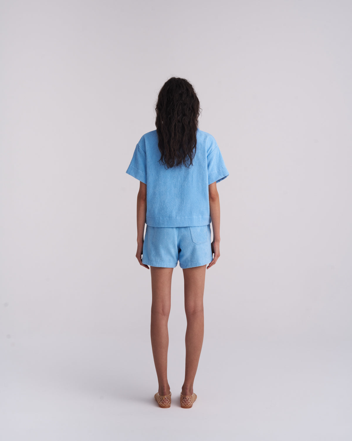 Woman in a Coastal Blue organic cotton terry camp-collar shirt and matching terry shorts, back view